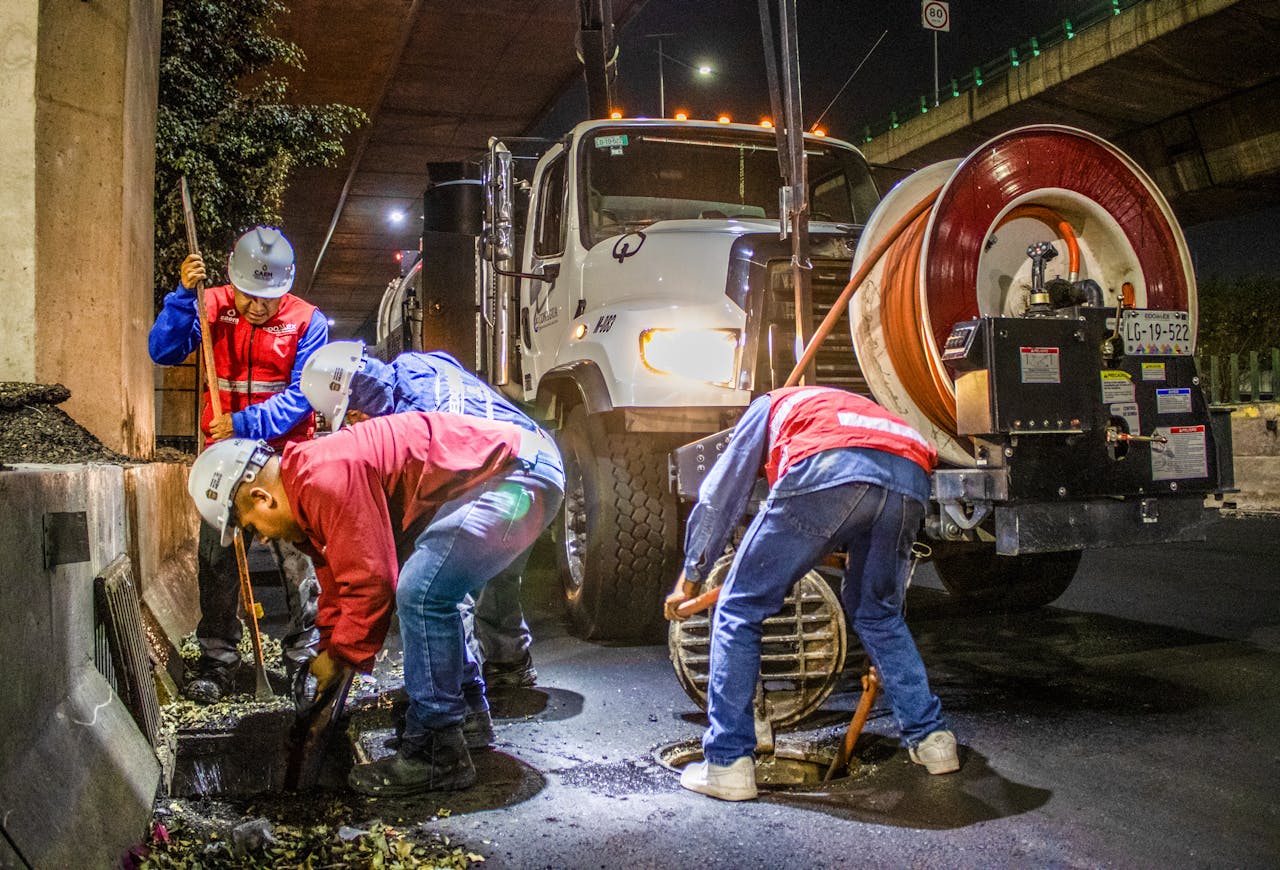 Street workers cleaning drainage at night with machinery under a bridge.